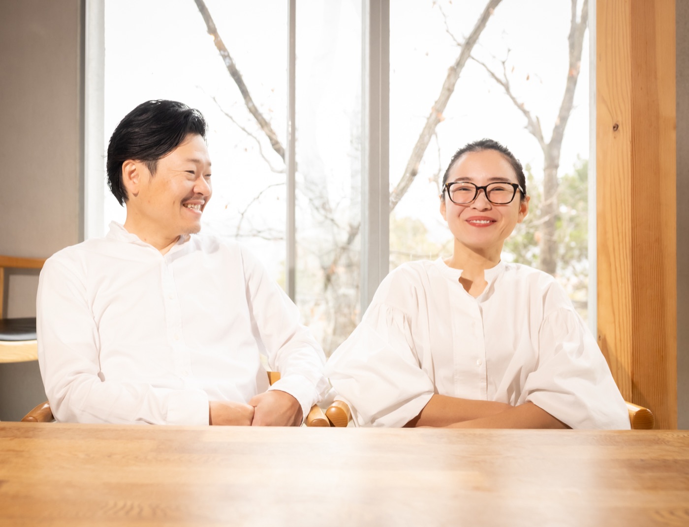窓辺の木のテーブルに並ぶHIROKIとASAKO。HIROKIがとなりのASAKOに目を向けている
